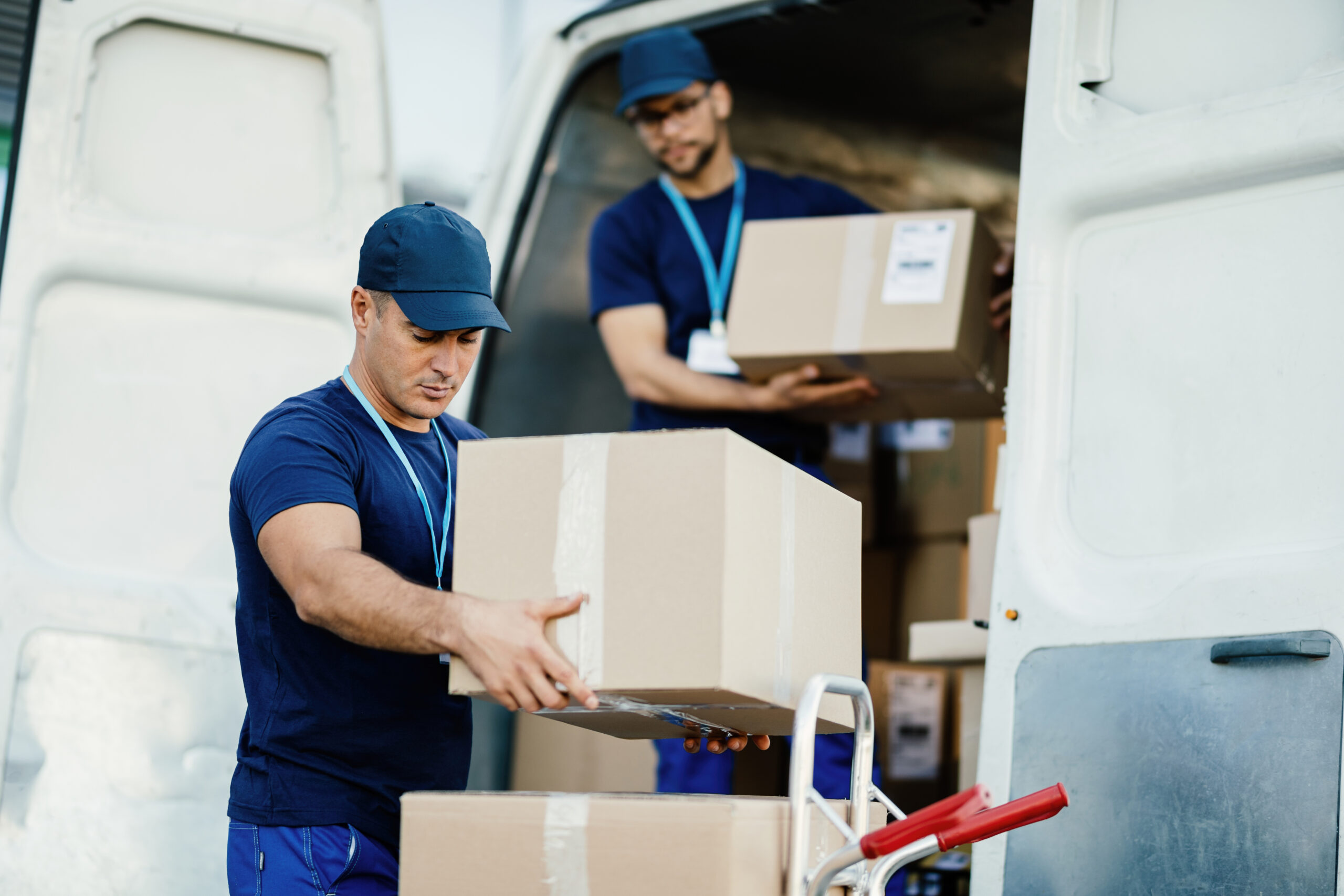 Young courier and his colleague unloading cardboard boxes from delivery van.